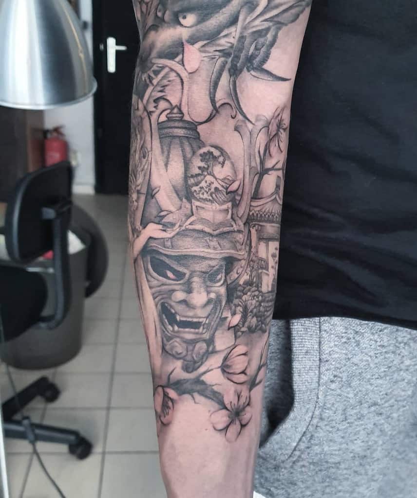 Intricate black and gray forearm tattoo featuring a fierce samurai mask, cherry blossoms, and Japanese architectural elements