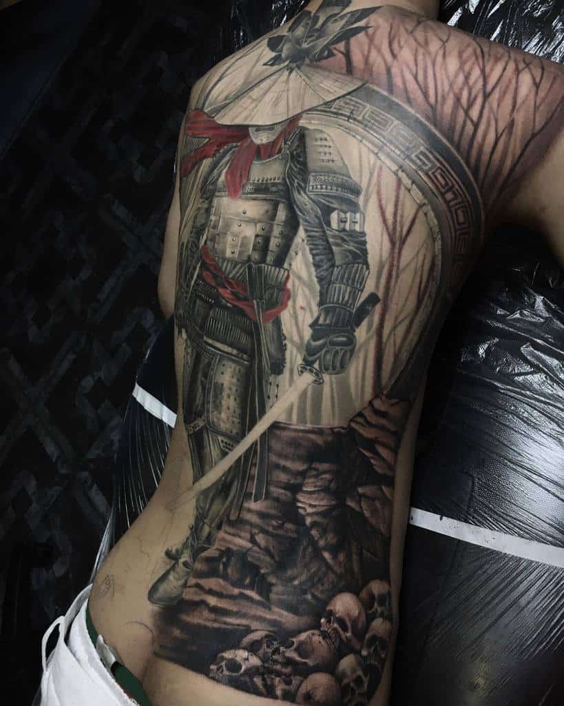 Stunning full-back tattoo of a Ronin in intricate armor with a red sash, standing amidst a forest and skull-covered terrain