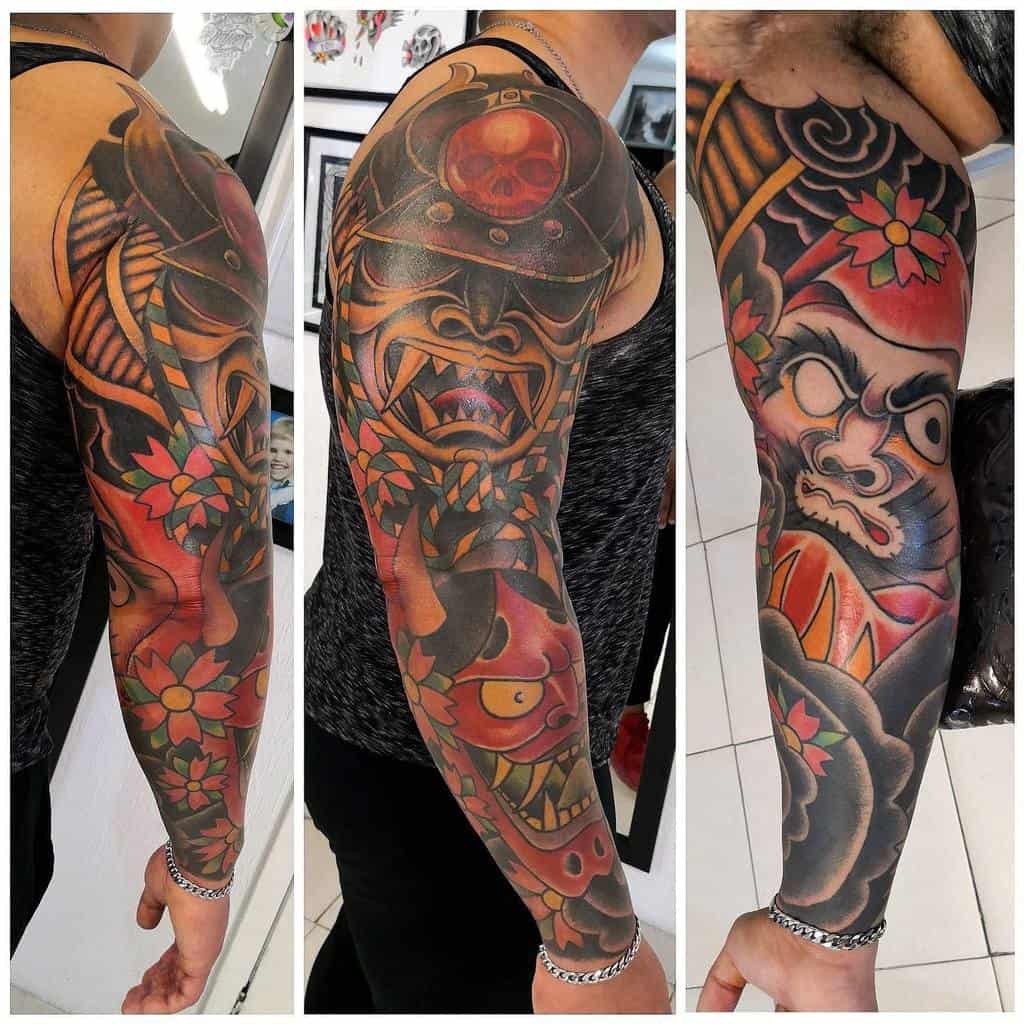 Vibrant full-sleeve tattoo showcasing a fierce samurai mask, bold red and orange tones, floral accents, and Japanese-inspired designs