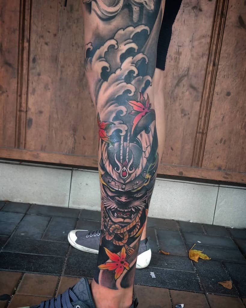 Colorful leg tattoo featuring a fierce samurai mask with intricate detailing, surrounded by swirling clouds and vibrant red maple leaves, blending traditional and modern Japanese art styles