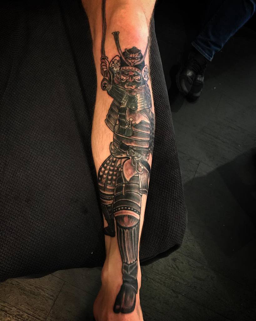 Leg tattoo features a striking samurai design, showcasing a bold and traditional Japanese warrior figure, emphasizing strength and honor with meticulous black and grey shading