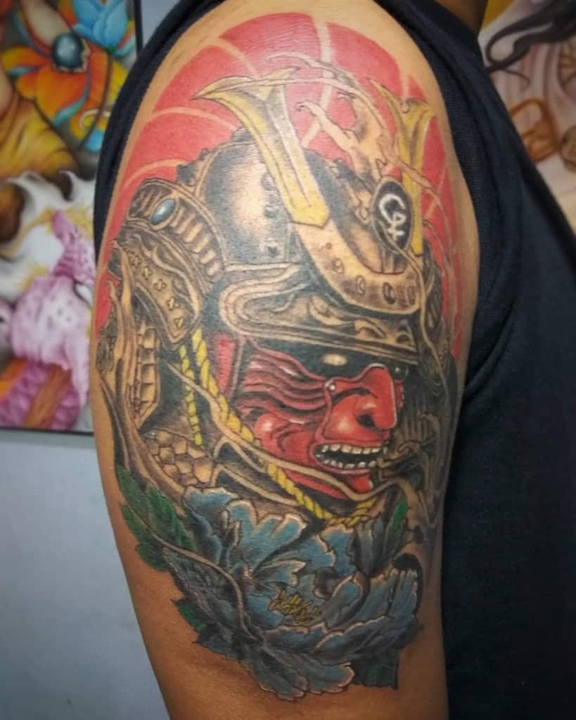 Vibrant samurai tattoo featuring a bold red mask, intricate armor details, a sunburst backdrop, and colorful peony flowers