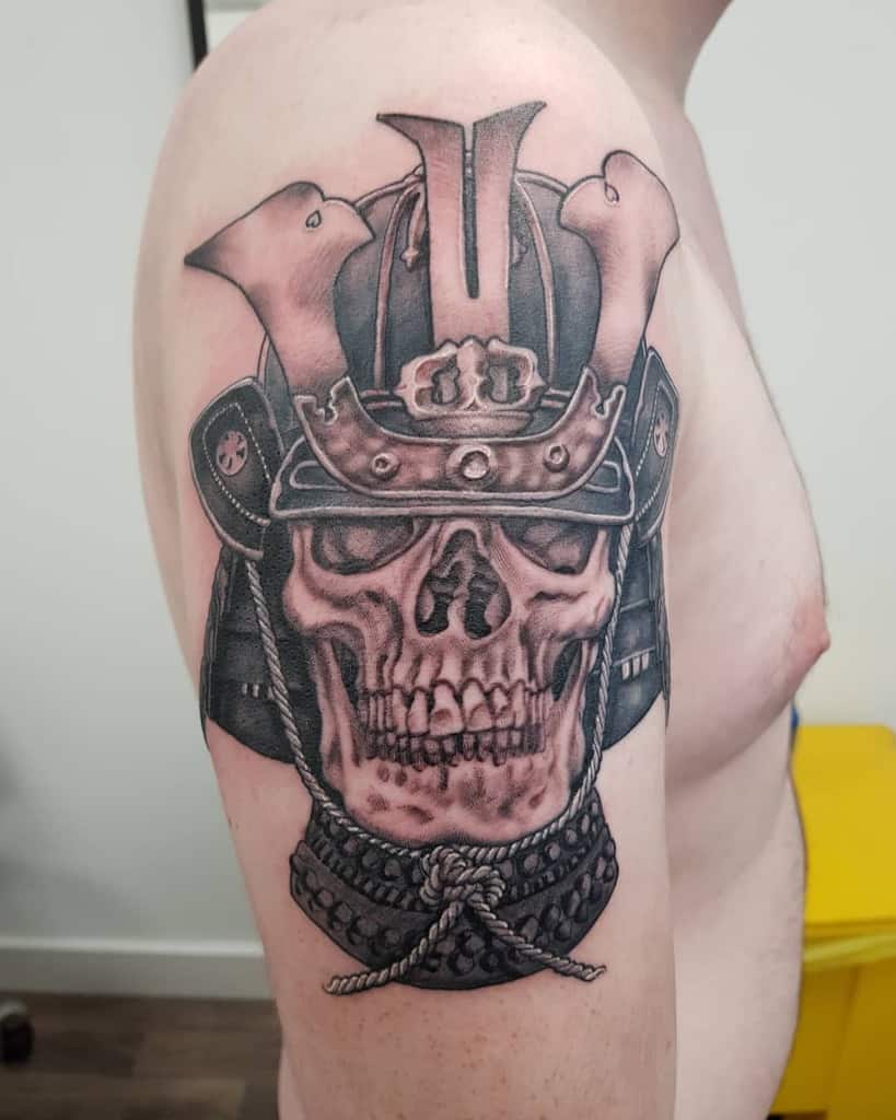 Striking black-and-gray tattoo of a skull wearing a samurai helmet, with detailed armor and a rope accent, emphasizing bold contrast and shading