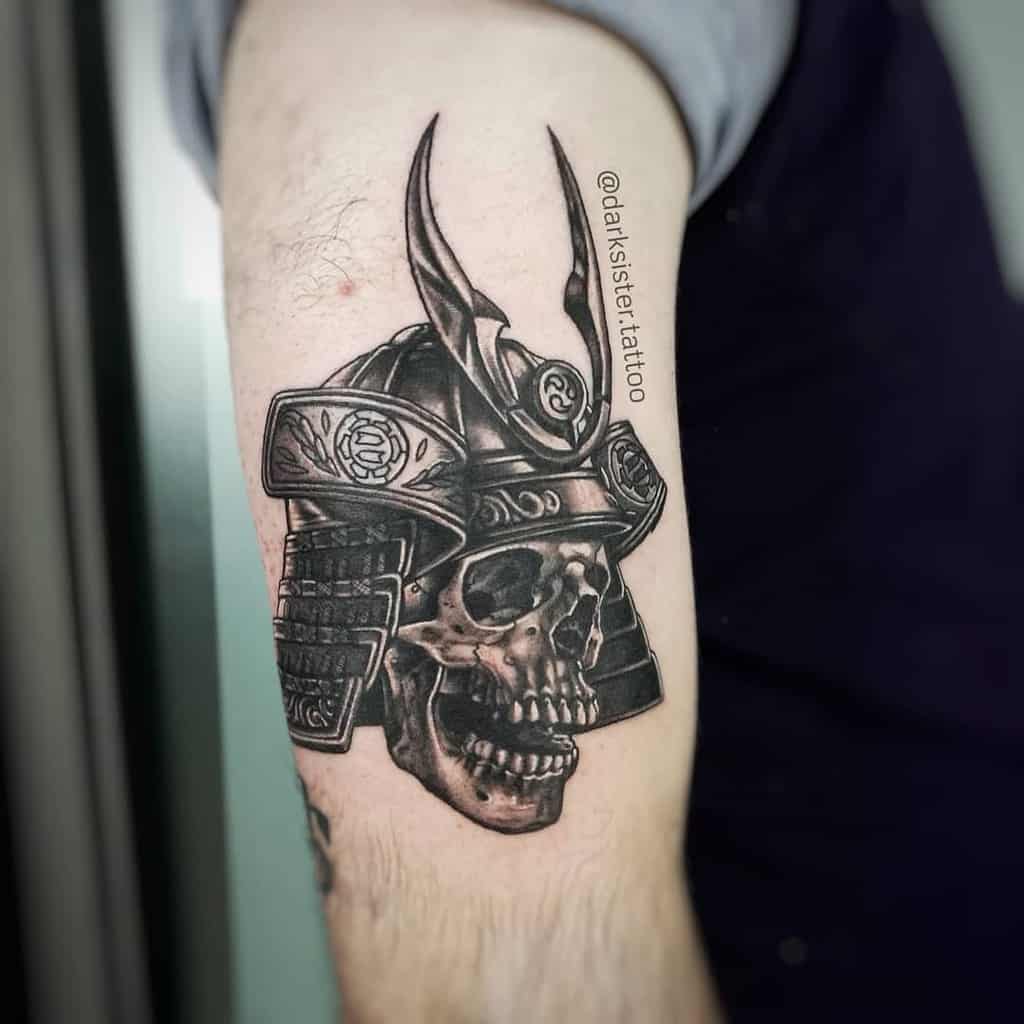 Intricate black-and-gray tattoo featuring a samurai skull helmet, with detailed textures on the armor and dramatic shading to highlight its fierce design