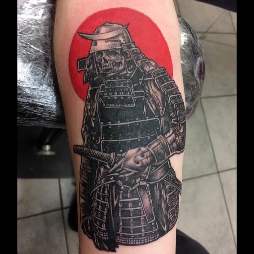 A striking black-and-gray tattoo of a samurai skull with intricate armor, standing proudly with a katana, set against a bold red sun backdrop