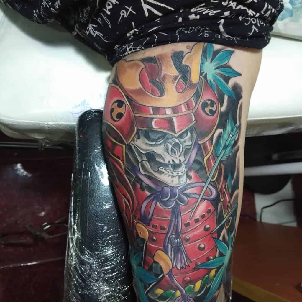 Vivid tattoo of a samurai skull adorned with colorful armor, surrounded by bamboo leaves and an arrow, emphasizing bold hues and dynamic shading