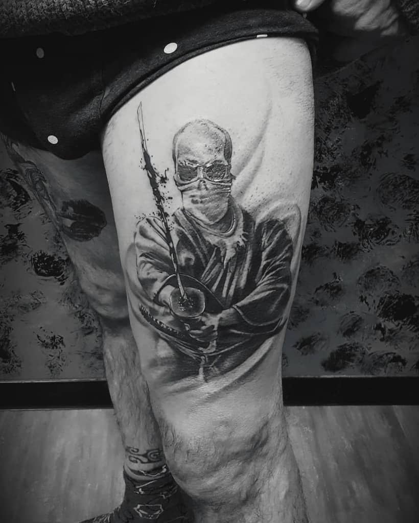Black and gray Ronin-inspired tattoo on a thigh, featuring a masked warrior holding a katana, detailed with ink splatter effects