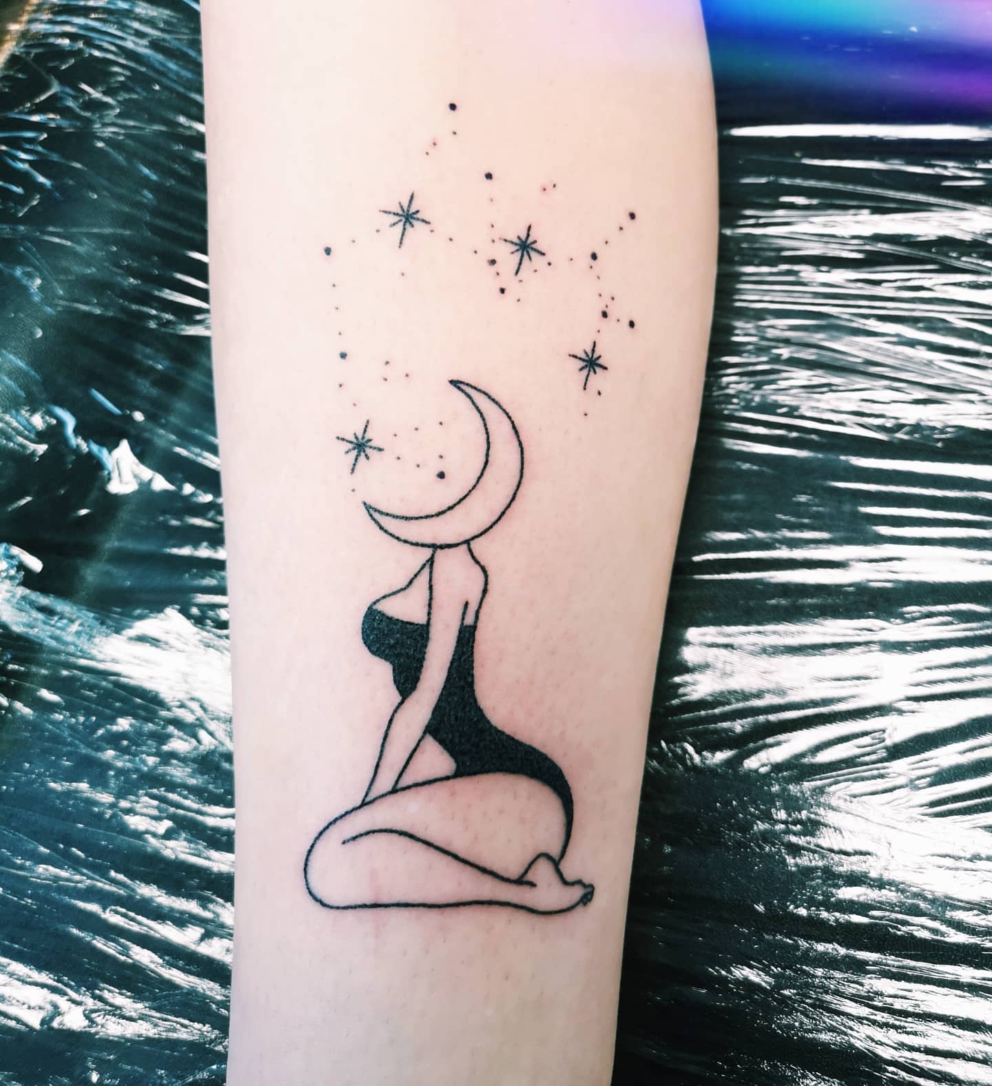 Tattoo of a sitting figure with a crescent moon head and stars above on an arm