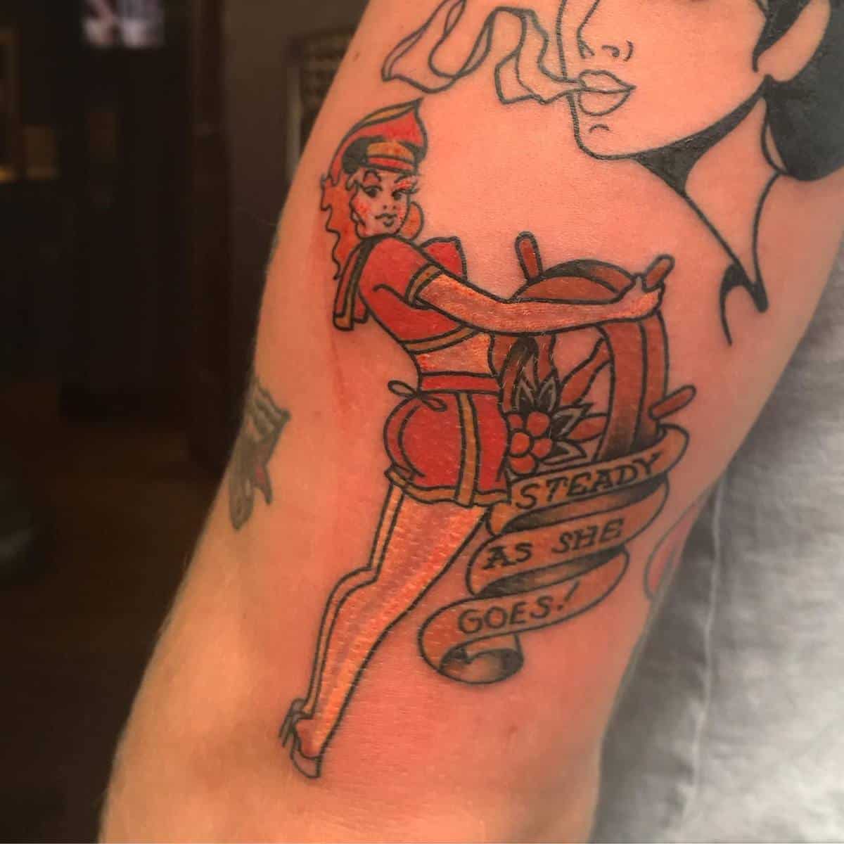 Tattoo of a pin-up girl in sailor attire with a "Steady As She Goes!" banner on a forearm