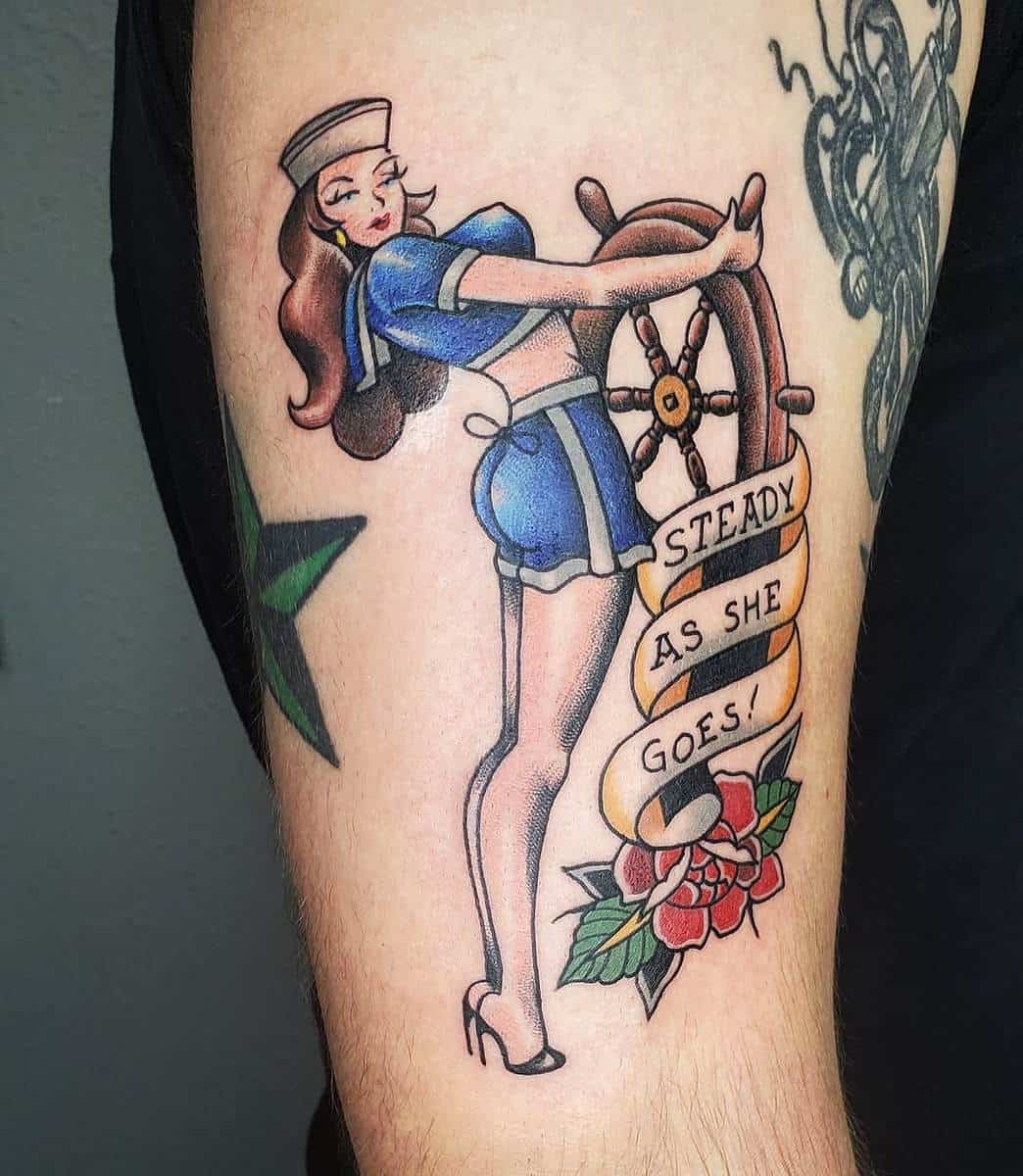 Tattoo of a sailor girl steering a ship's wheel with "Steady As She Goes!" banner and a rose below