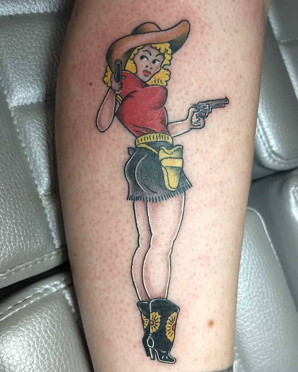 Tattoo of a vintage-style cowgirl with blonde hair, red top, black shorts, and holding two guns on a leg