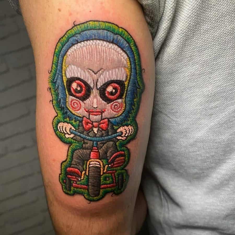 Patch-style embroidery tattoo of the Saw movie puppet, featuring red spiral cheeks, a bow tie, and a tricycle in a stitched effect.