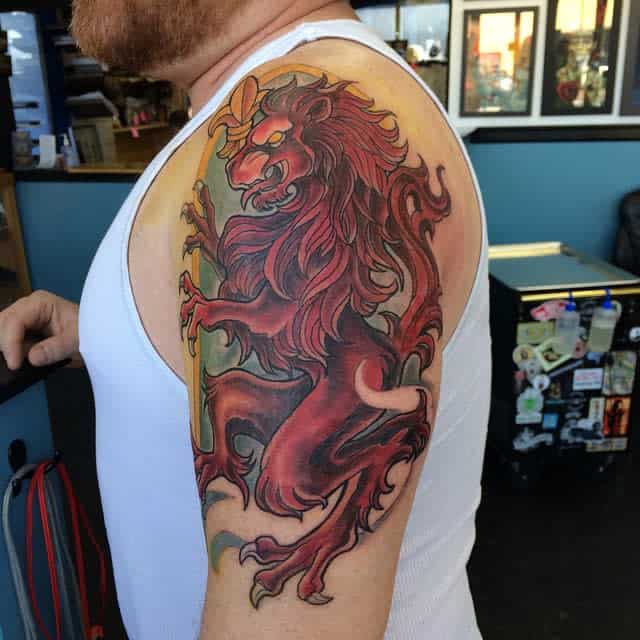 Bold tattoo of a red, heraldic Scottish lion rampant with a fierce expression, gripping a golden staff with green background