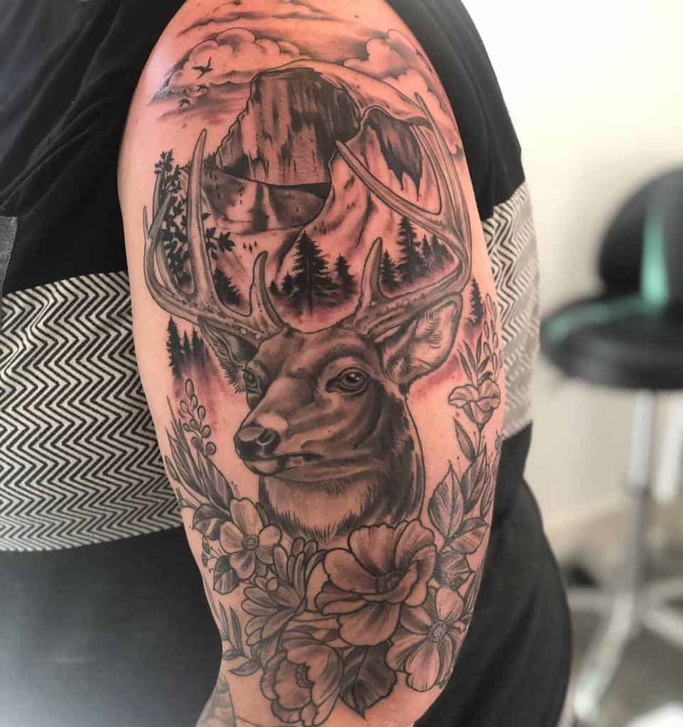 Arm tattoo of a Scottish stag with antlers, surrounded by flowers, leaves, trees, birds and high mountains under a cloudy sky