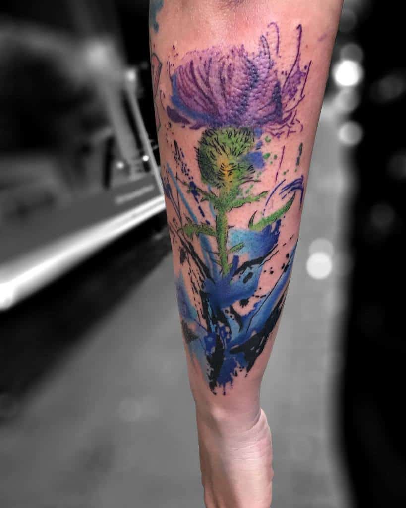 Dramatic watercolor-style tattoo of a Scottish thistle, with vivid blue and purple hues