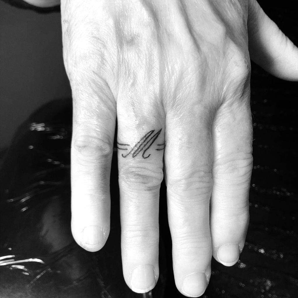 A minimalist ring tattoo featuring the letter "M" with horizontal lines on either side