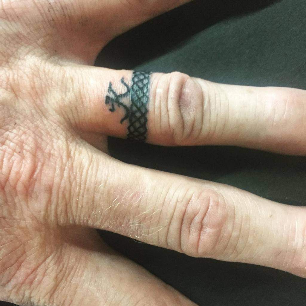 A bold black tattoo featuring a lace-like pattern around the finger, with a small symbol 'K'