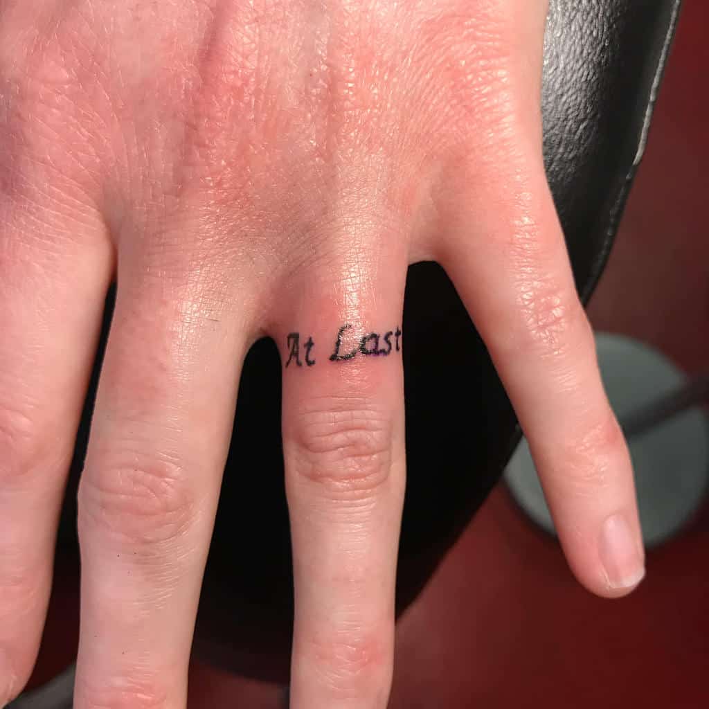 A ring tattoo on the finger featuring the phrase "At Last" in elegant cursive font