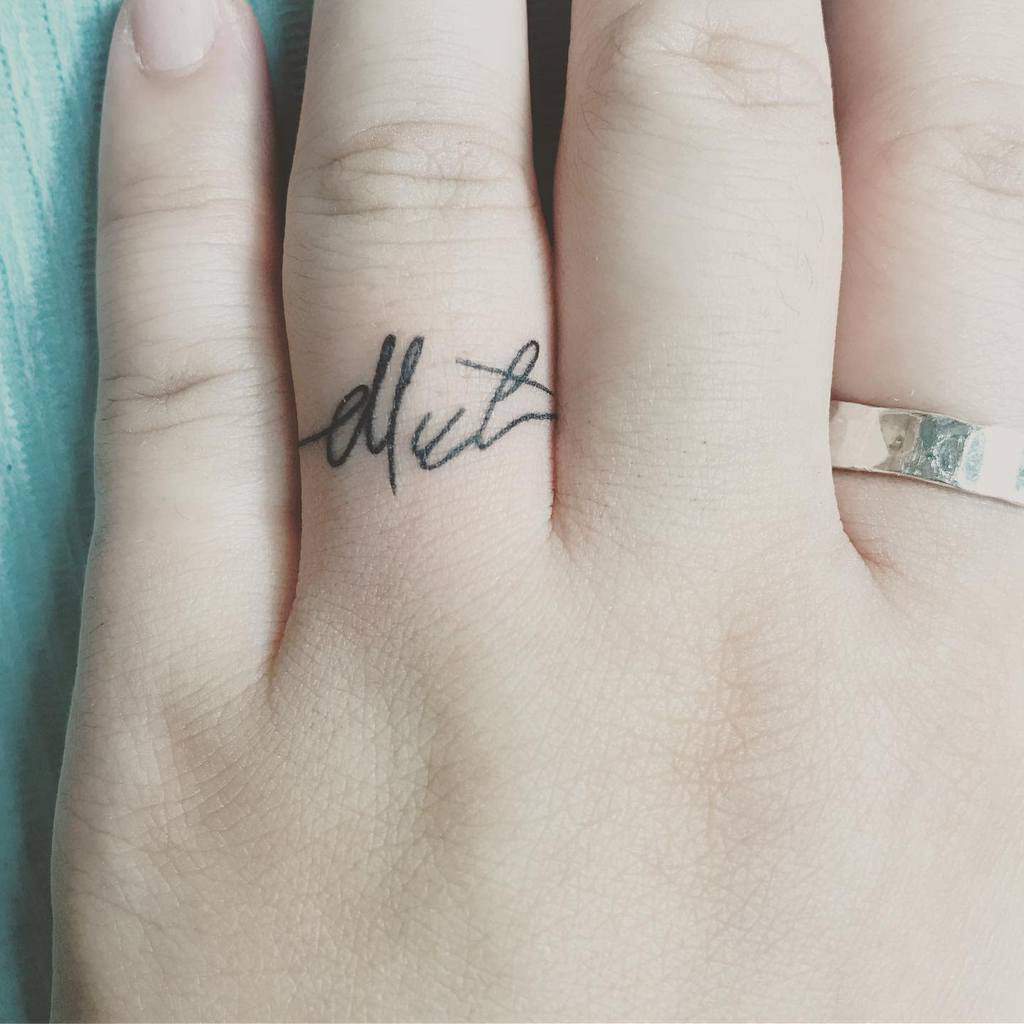 A ring tattoo on the finger featuring a cursive signature-style word "dlut," in bold black ink