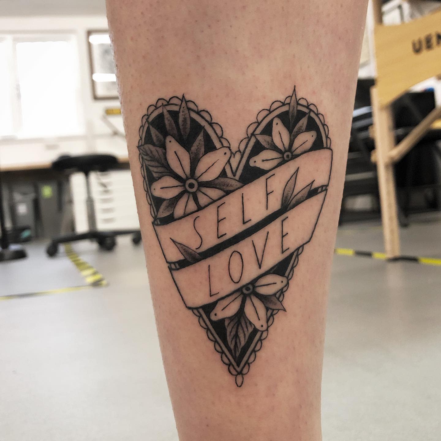 Heart-shaped tattoo with flowers and a ribbon that reads "SELF LOVE" on a leg