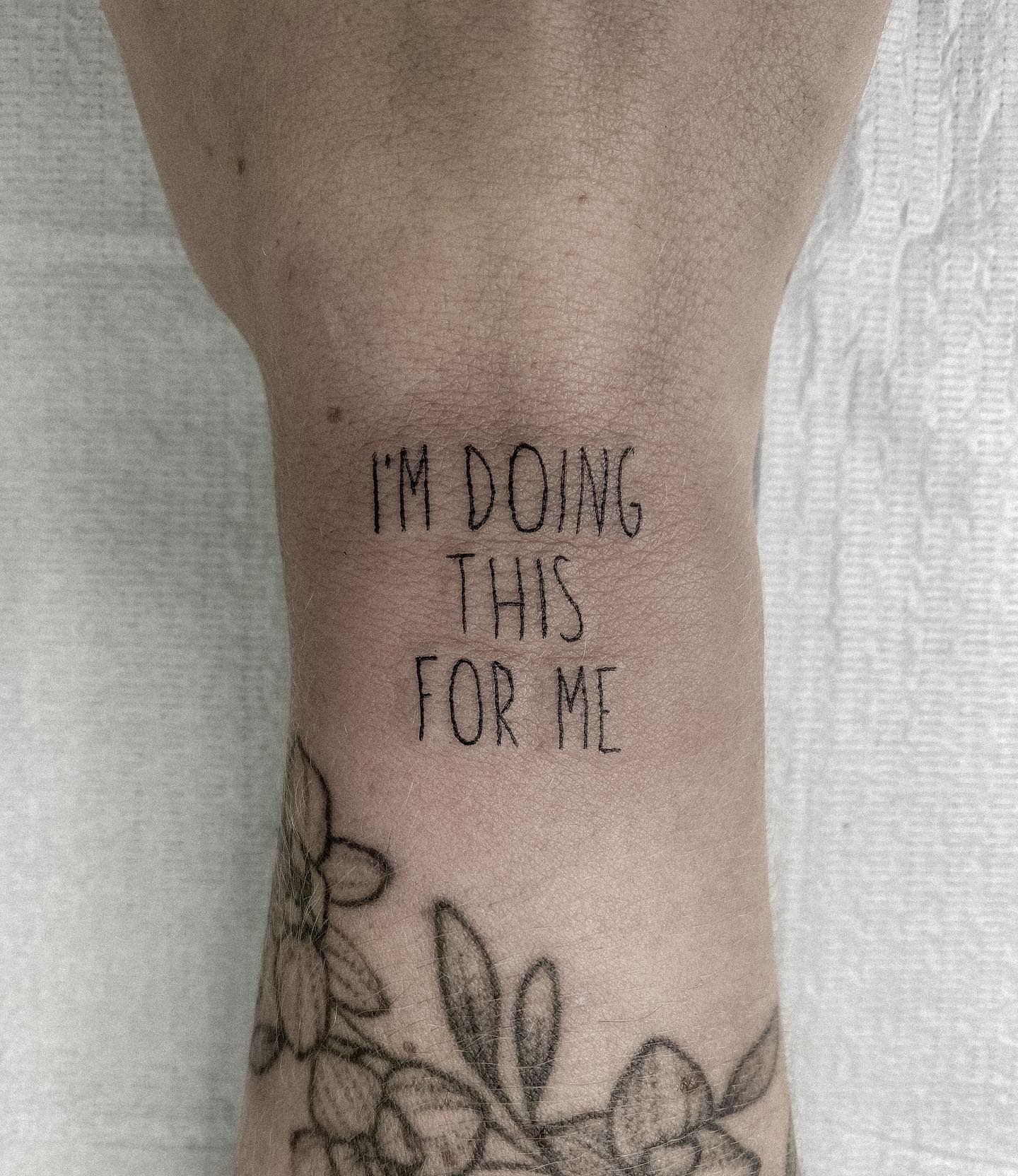 Tattoo on an arm reading "I'M DOING THIS FOR ME" above a floral tattoo