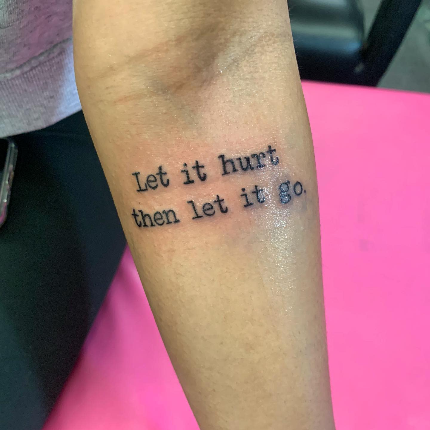 Tattoo on forearm with text "Let it hurt then let it go" on pink background