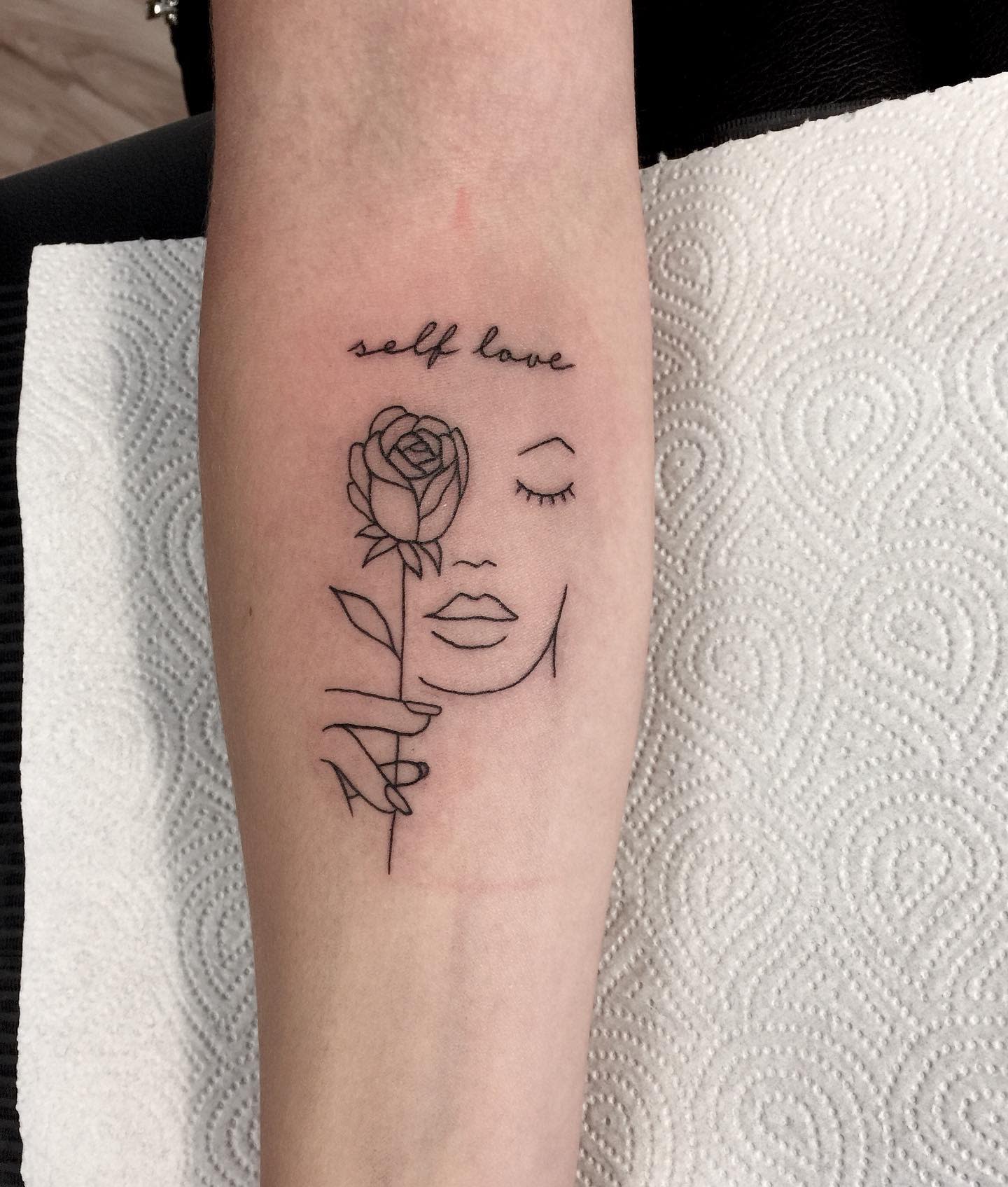 Line drawing tattoo of a face with a rose, captioned "self love," on a forearm