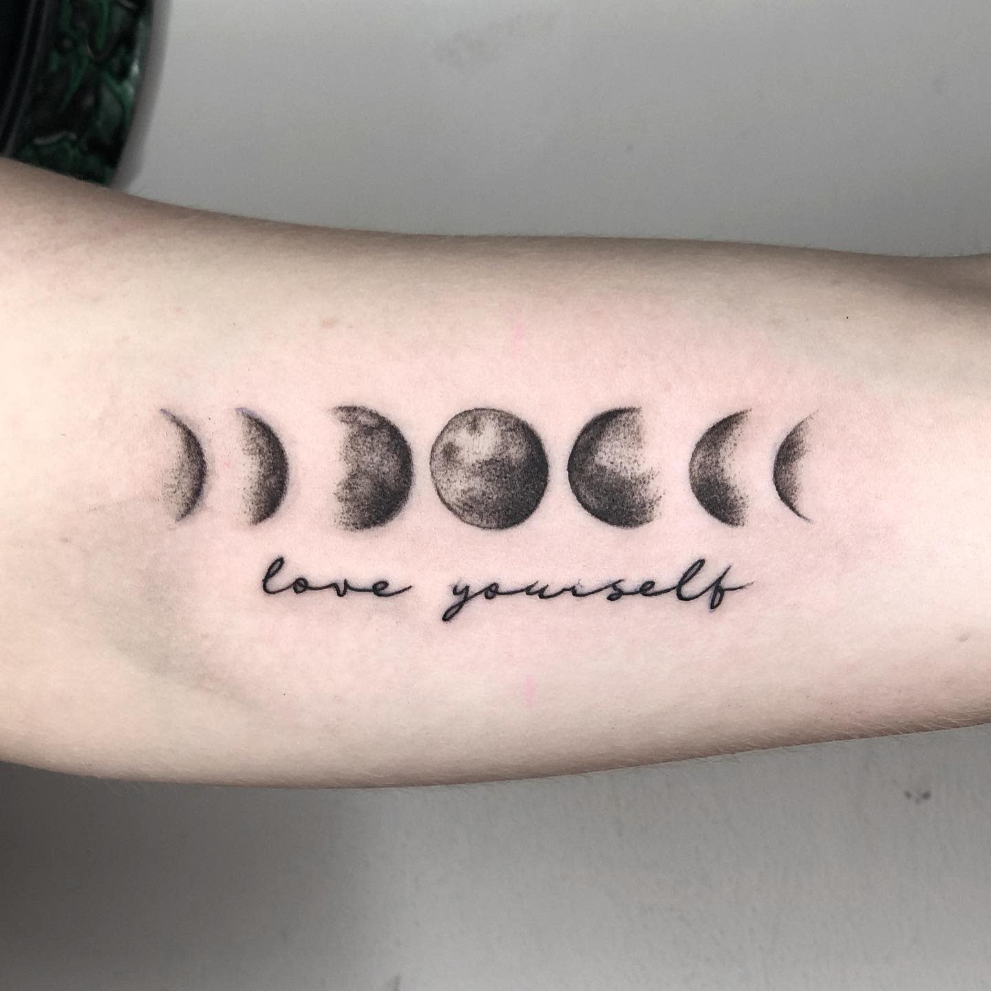 Tattoo of moon phases with “love yourself” text below located on inner forearm
