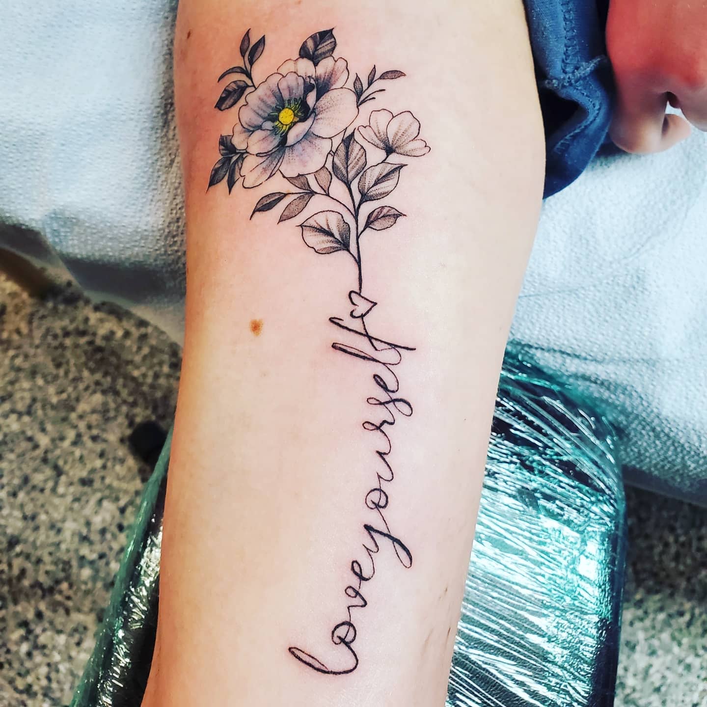 Tattoo of flowers with cursive text spelling "love yourself" on someone's arm