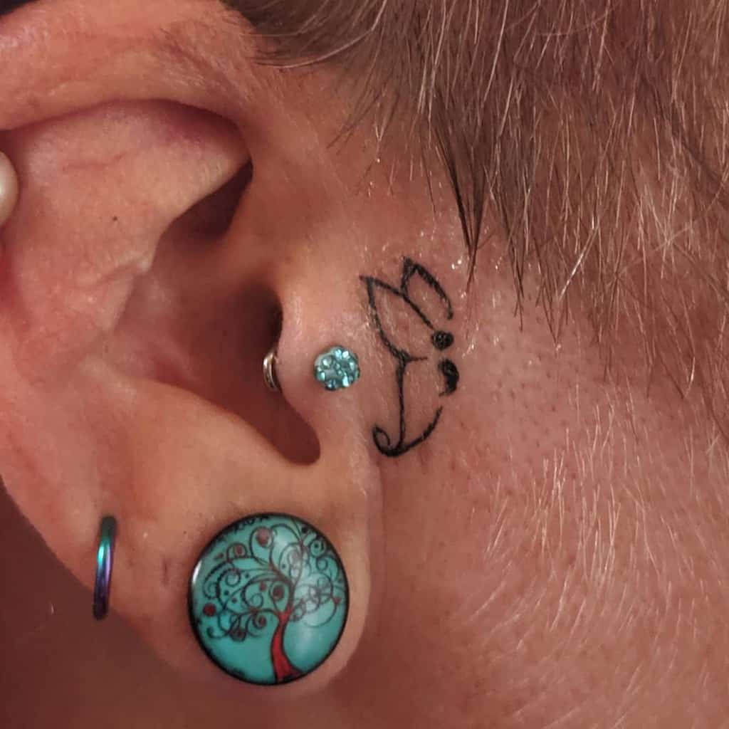 A tiny and minimalist black semicolon butterfly tattoo placed behind the ear, symbolizing inner strength, mental health awareness, and subtle beauty