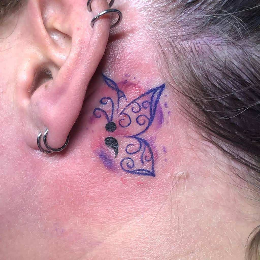 A delicate semicolon butterfly tattoo behind the ear with intricate swirls and soft purple shading, symbolizing transformation, mental health awareness, and personal resilience