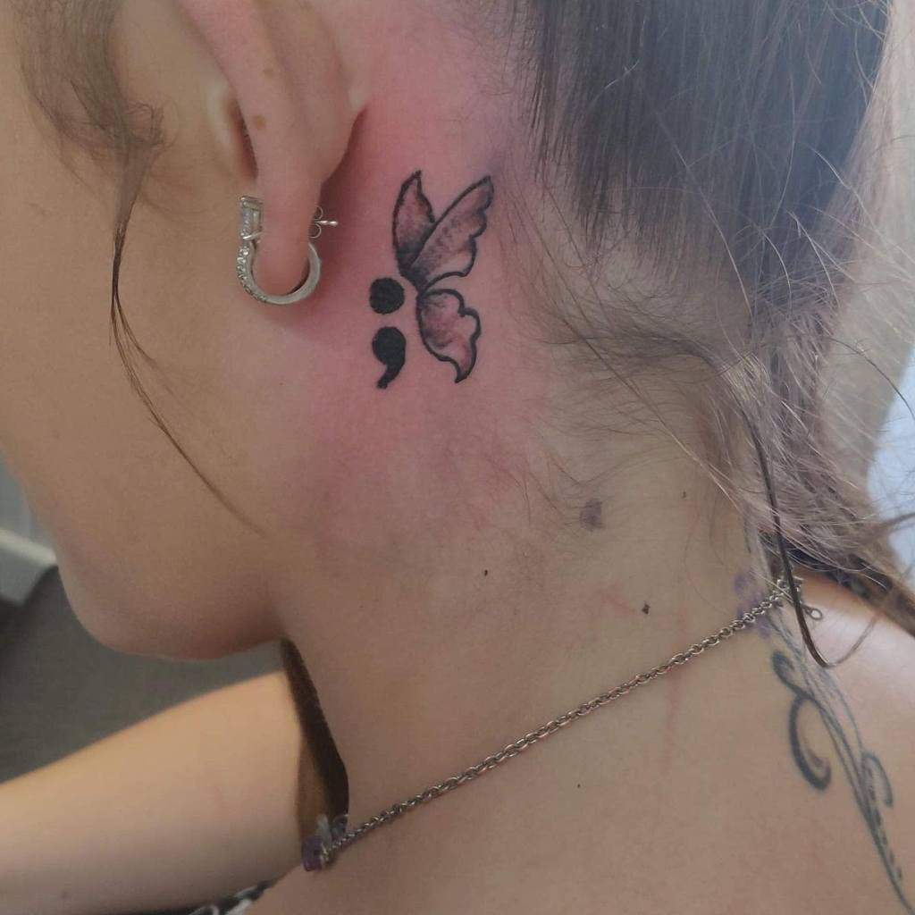 A small, minimalist semicolon butterfly tattoo behind the ear, representing strength, mental health awareness, and overcoming personal struggles