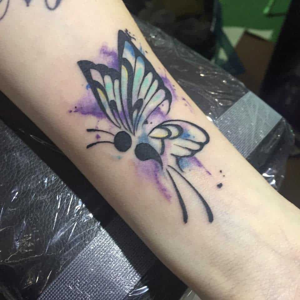 A striking semicolon butterfly tattoo on the forearm with black and pastel blue wings, accented by purple watercolor splashes, symbolizing mental health awareness, personal growth, and the beauty of transformation
