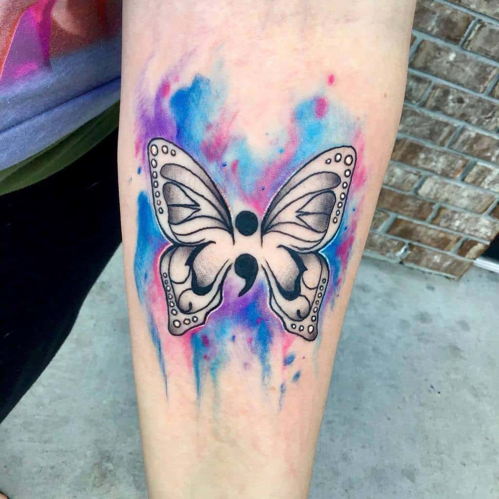 A black and white semicolon butterfly tattoo on the forearm with colorful blue and pink watercolor splashes, symbolizing mental health awareness, personal growth, and the vibrant journey of transformation