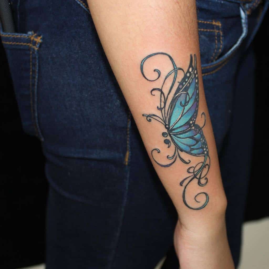 A stunning blue semicolon butterfly tattoo on the forearm, accented with elegant swirls and dotwork, symbolizing transformation, hope, and mental health awareness