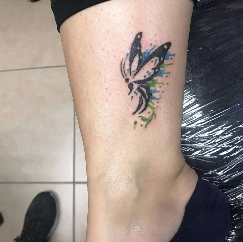 Abstract black butterfly tattoo with vibrant watercolor splashes in blue and green, creating a dynamic and artistic effect.