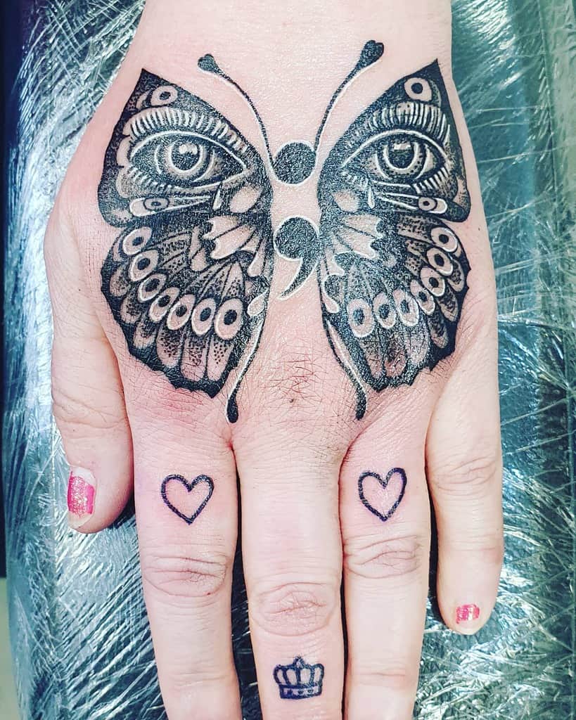 Intricate black and grey butterfly tattoo with realistic eyes and a semicolon design on the hand, symbolizing awareness and transformation.