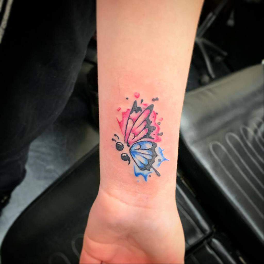 A small watercolor butterfly tattoo with pink and blue wings and a semicolon body on the wrist, symbolizing hope and mental health awareness