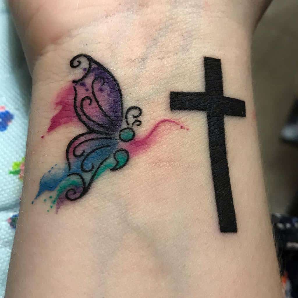 A semicolon butterfly tattoo with colorful swirls next to a bold black cross on the wrist, symbolizing faith, hope, and mental health awareness