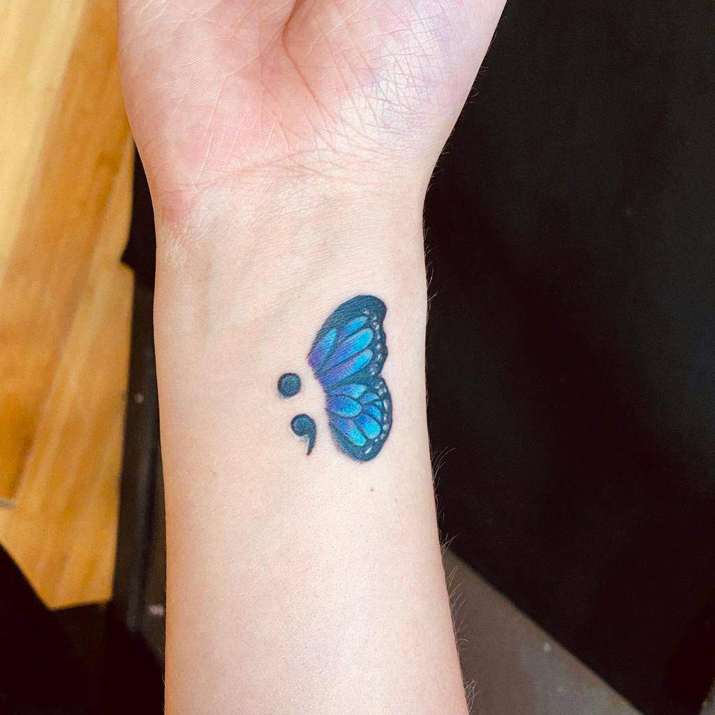 A small blue and purple butterfly tattoo with a semicolon body on the wrist, symbolizing mental health awareness and personal strength