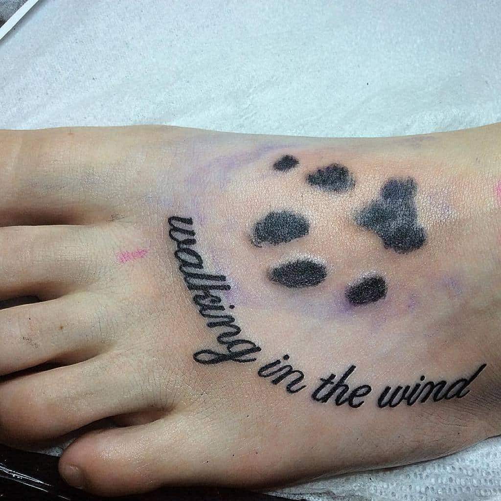 Tattoo on foot with cat paw prints and the text "walking in the wind" in cursive