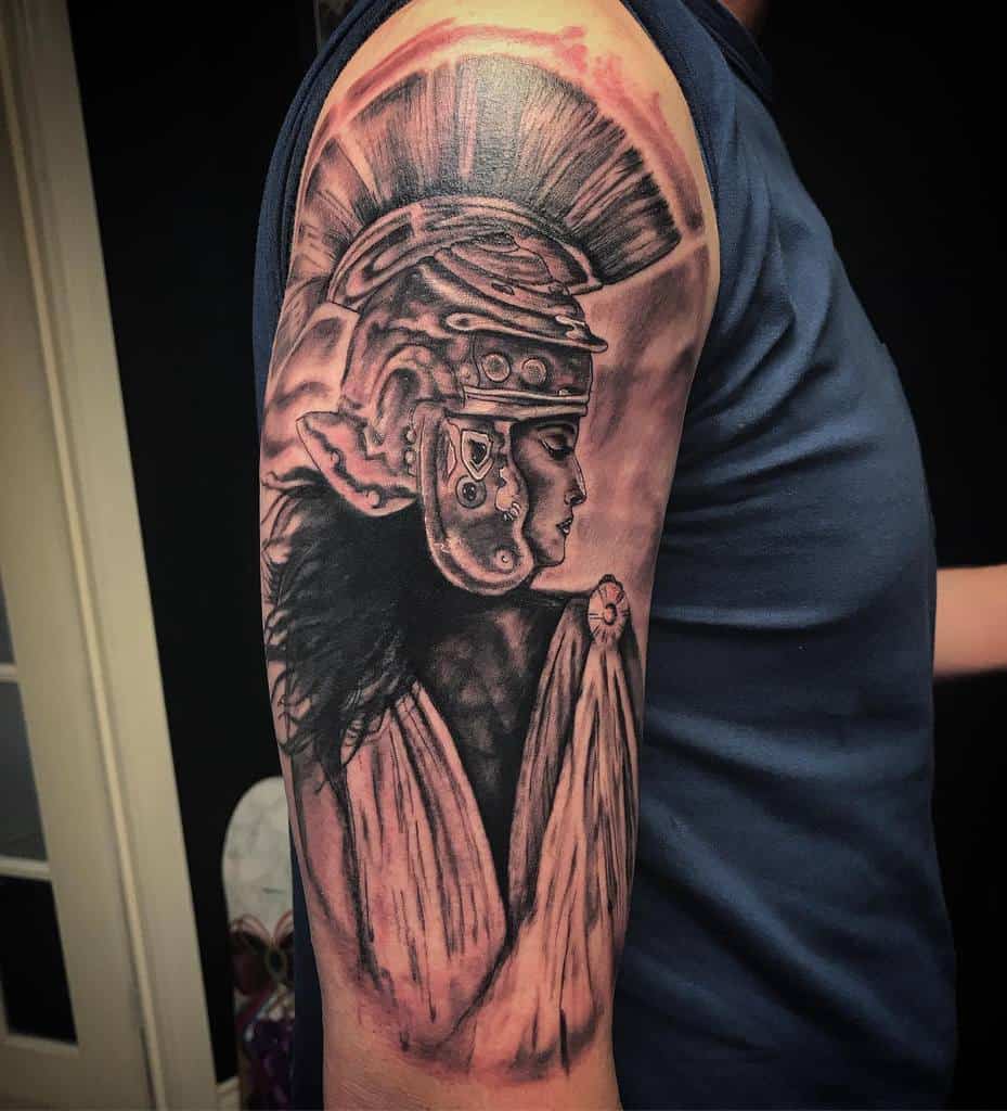 A black and grey half sleeve tattoo featuring a detailed portrait of a warrior wearing a Roman-style helmet, with realistic shading and bold contrast, covering the upper arm