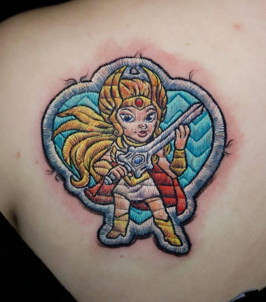 Embroidered patch-style tattoo of a warrior princess in gold armor, wielding a sword with a determined expression.