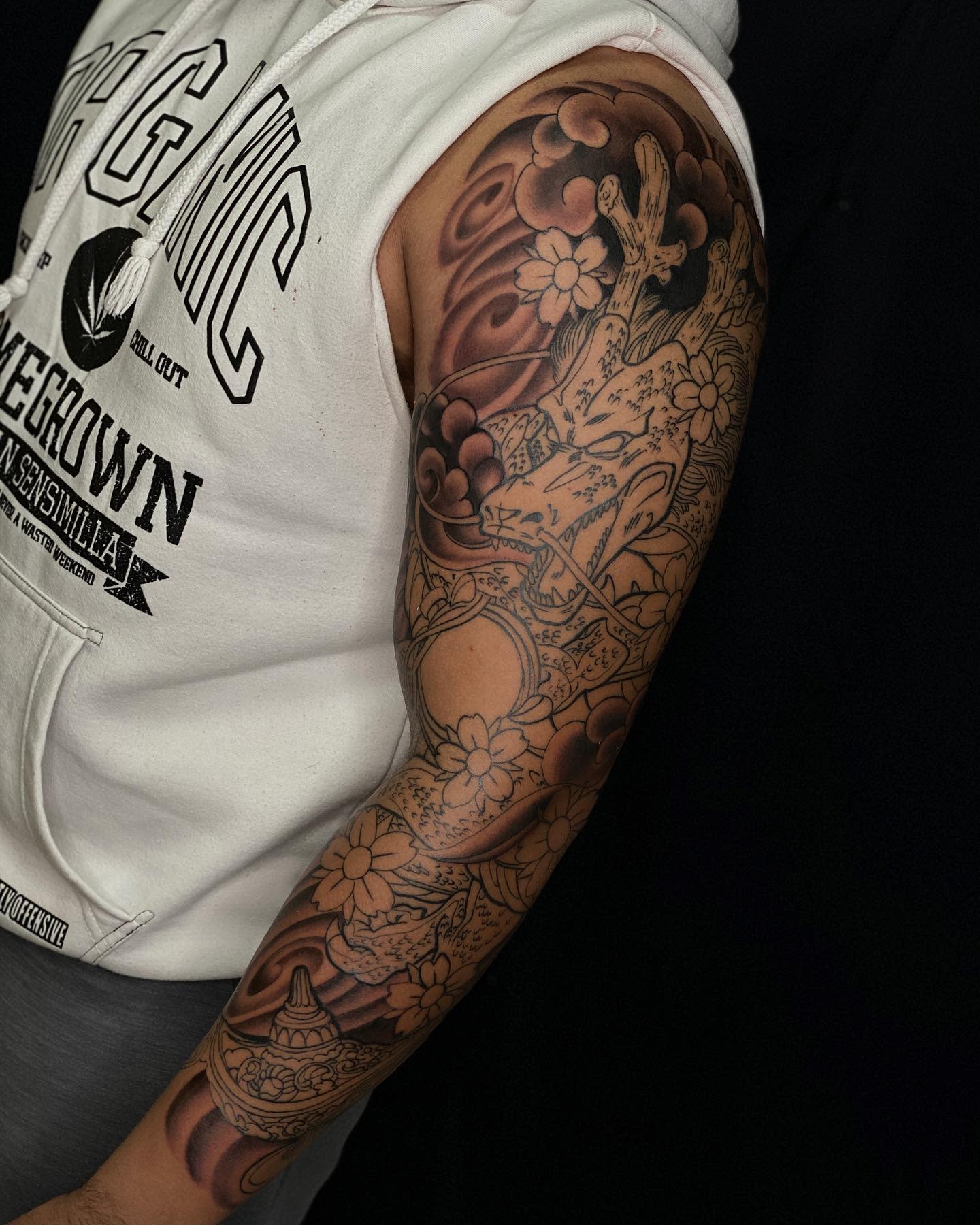 Complex sleeve tattoo showcasing Shenron intertwined with flowers and clouds, with portions shaded