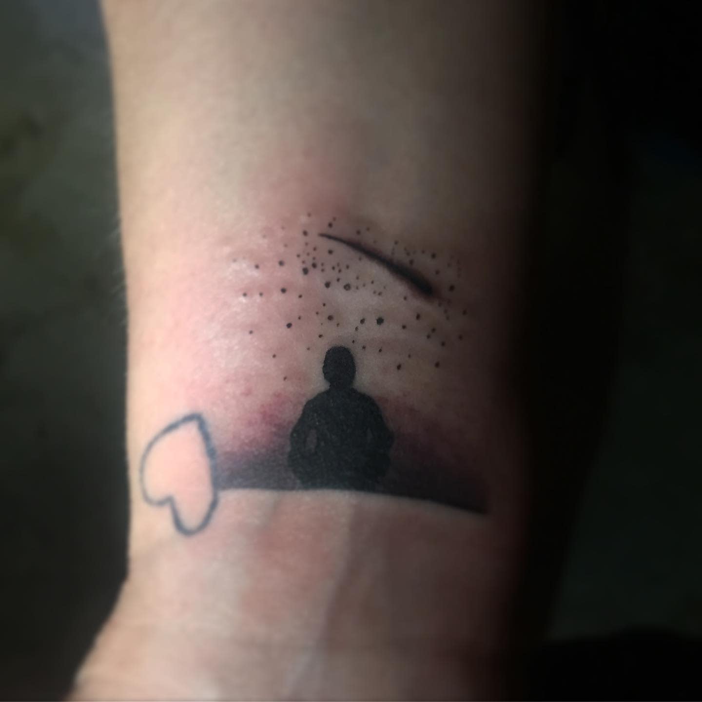Tattoo of someone sitting under a sky with stars and a shooting star next to a heart