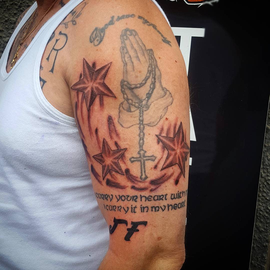 Tattooed arm with praying hands, a cross, stars, and a quote with decorative elements
