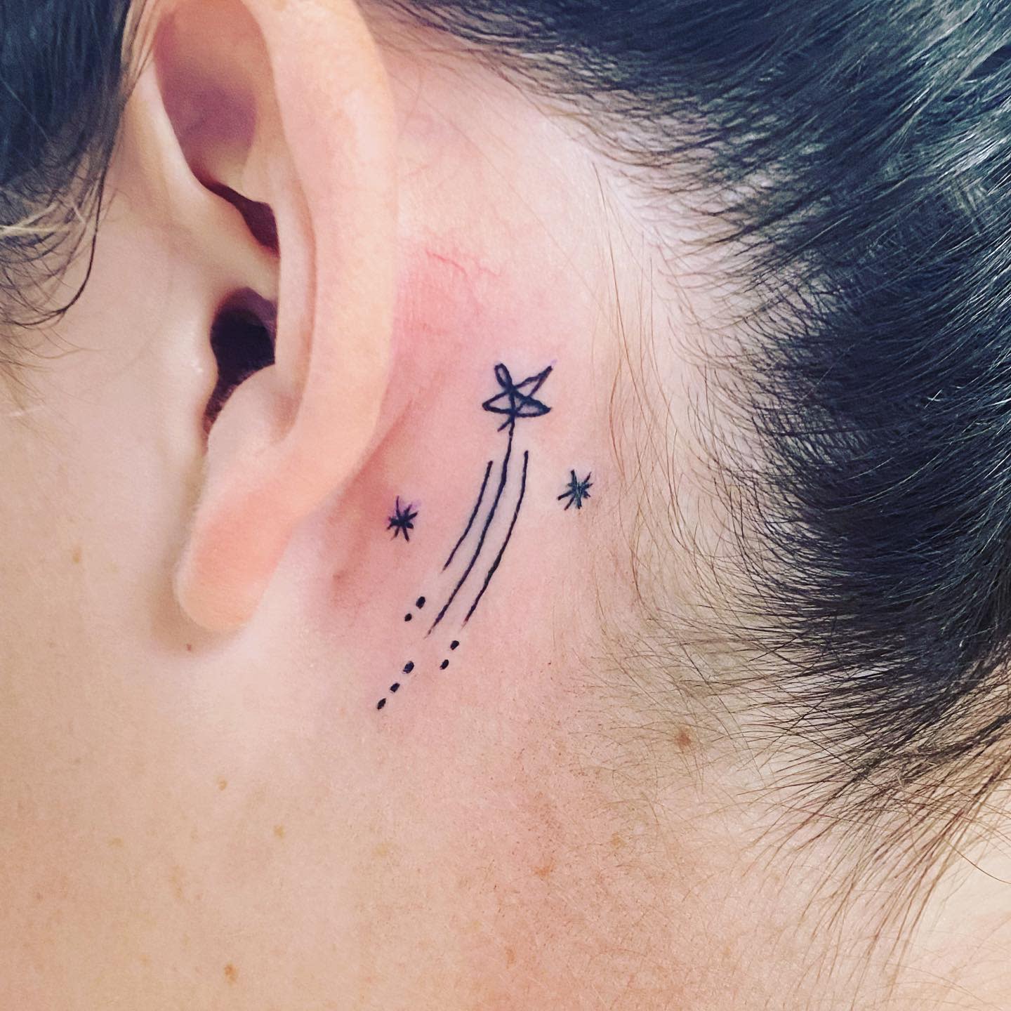 Tattoo of a shooting star near the ear with small stars surrounding it