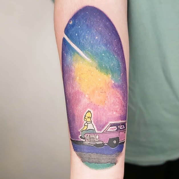 Tattoo of Homer Simpson sitting on a car roof, gazing at a colorful galaxy on the forearm