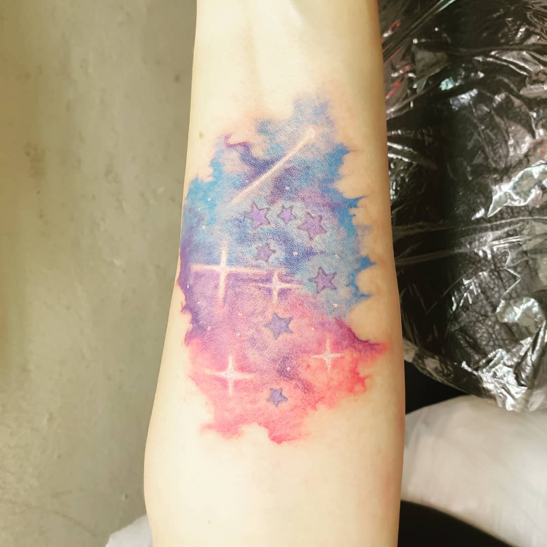 Watercolor galaxy tattoo with stars and glowing accents on an inner forearm