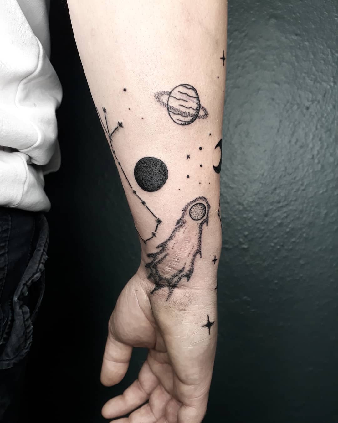 Forearm tattoo of planets, a constellation, and a space-themed rocket against a dark backdrop
